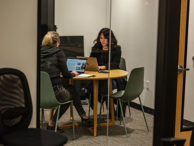 Two women collaborating at a round table in a modern office setting, engaged in discussion with a laptop and notes, reflecting the creative and community-focused environment of Murmur Creative.